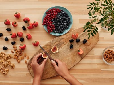 Mãos preparando frutas e oleaginosas sobre uma tábua de madeira, cercadas por ingredientes vegetais frescos