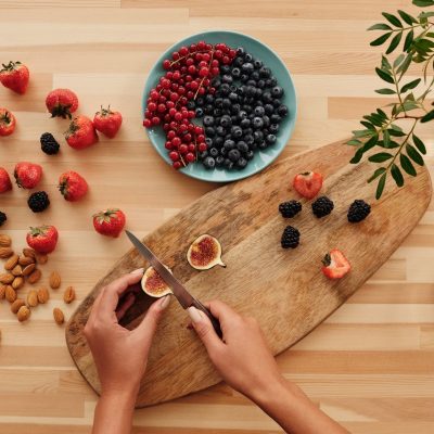 Mãos preparando frutas e oleaginosas sobre uma tábua de madeira, cercadas por ingredientes vegetais frescos