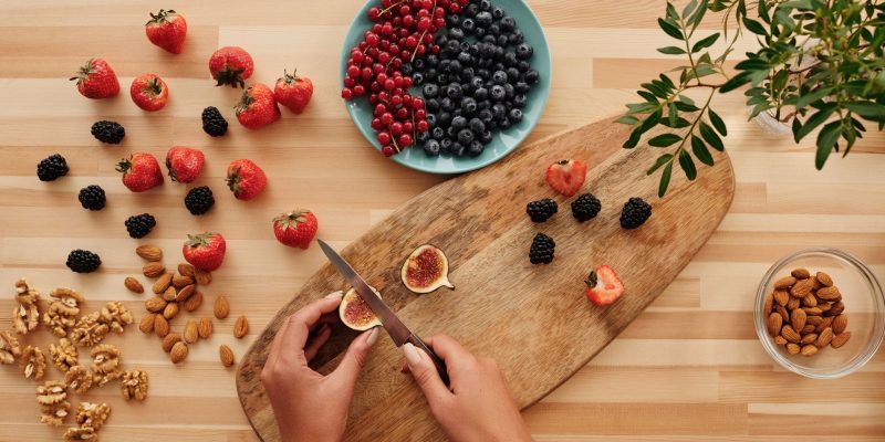 Mãos preparando frutas e oleaginosas sobre uma tábua de madeira, cercadas por ingredientes vegetais frescos