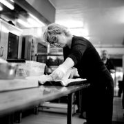 Chef concentrada prepara alimentos sobre uma bancada metálica em uma cozinha profissional.
