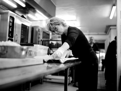 Chef concentrada prepara alimentos sobre uma bancada metálica em uma cozinha profissional.