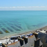 Vista aérea da costa de Cancale, na Bretanha, com viveiros de ostras, mar azul e um farol ao fundo.