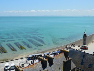 Vista aérea da costa de Cancale, na Bretanha, com viveiros de ostras, mar azul e um farol ao fundo.