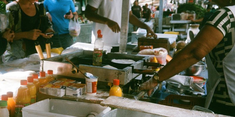 Público circula entre barracas de uma feira de comida em São Paulo, com vendedores e produtos expostos.